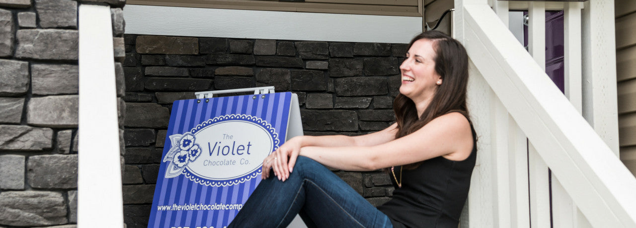 Rebecca sits in front of studio with sign of The Violet Chocolate Company click to learn more about her story